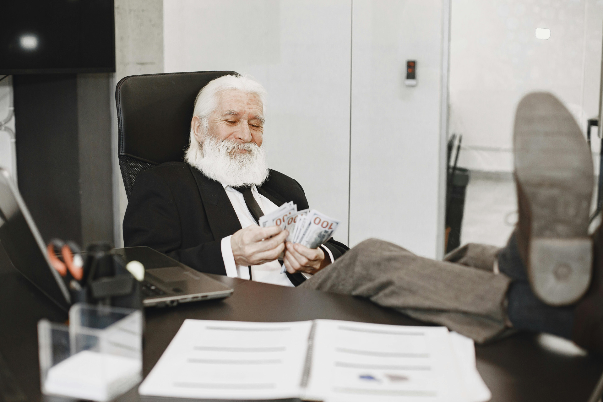 Elderly businessman relaxing at desk, counting money notes in a modern office setting.